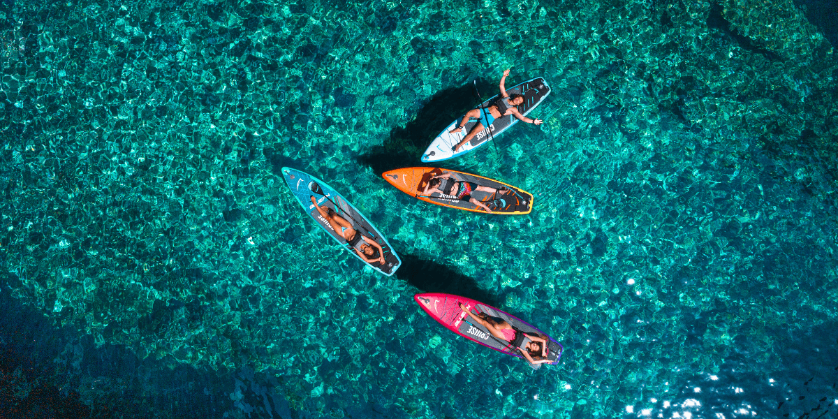 Four people paddle colourful stand-up paddleboards in clear turquoise water, viewed from above