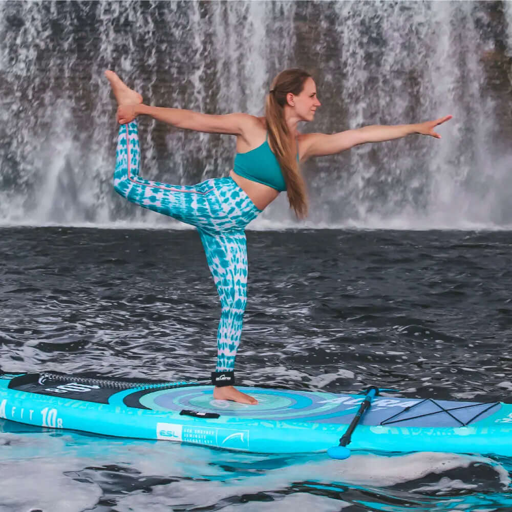 Mujer en ropa activa color teal haciendo yoga en una tabla de paddle frente a una cascada