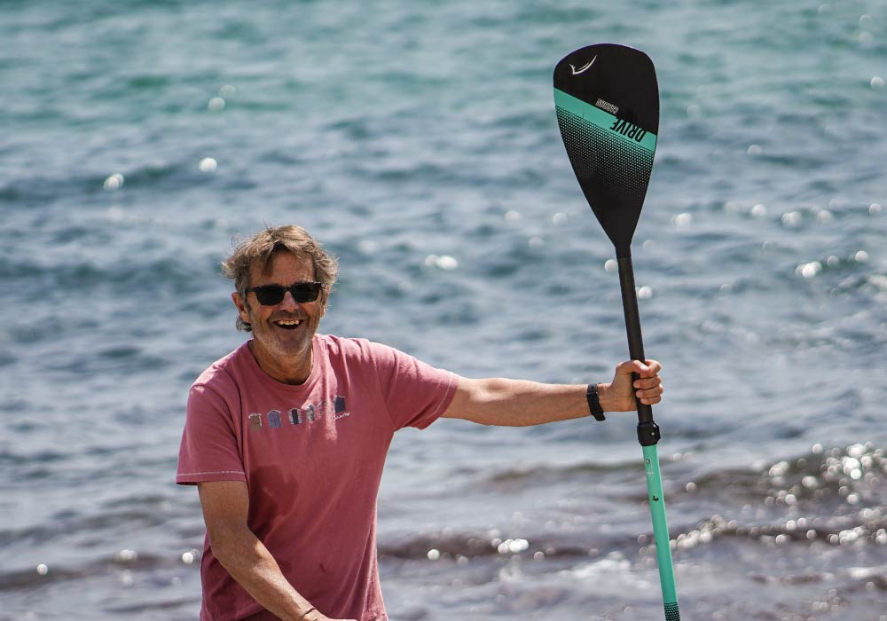 Man in a pink shirt holding a black and teal paddle near the water, smiling at the camera.