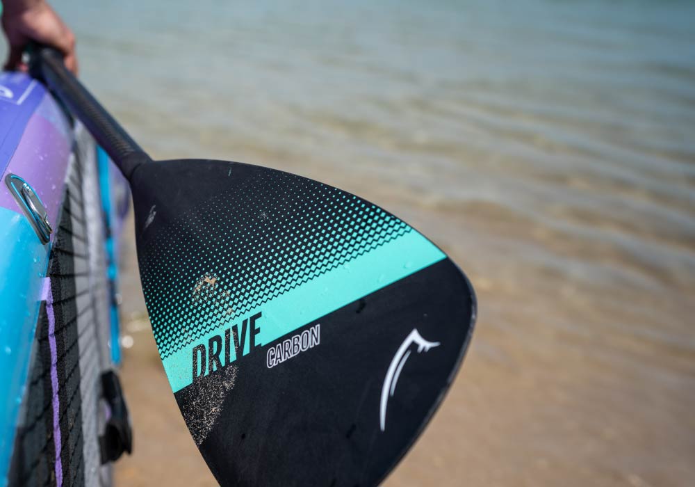 Close-up of a carbon paddle blade with 'DRIVE' text, held above water with a colorful paddleboard in background.