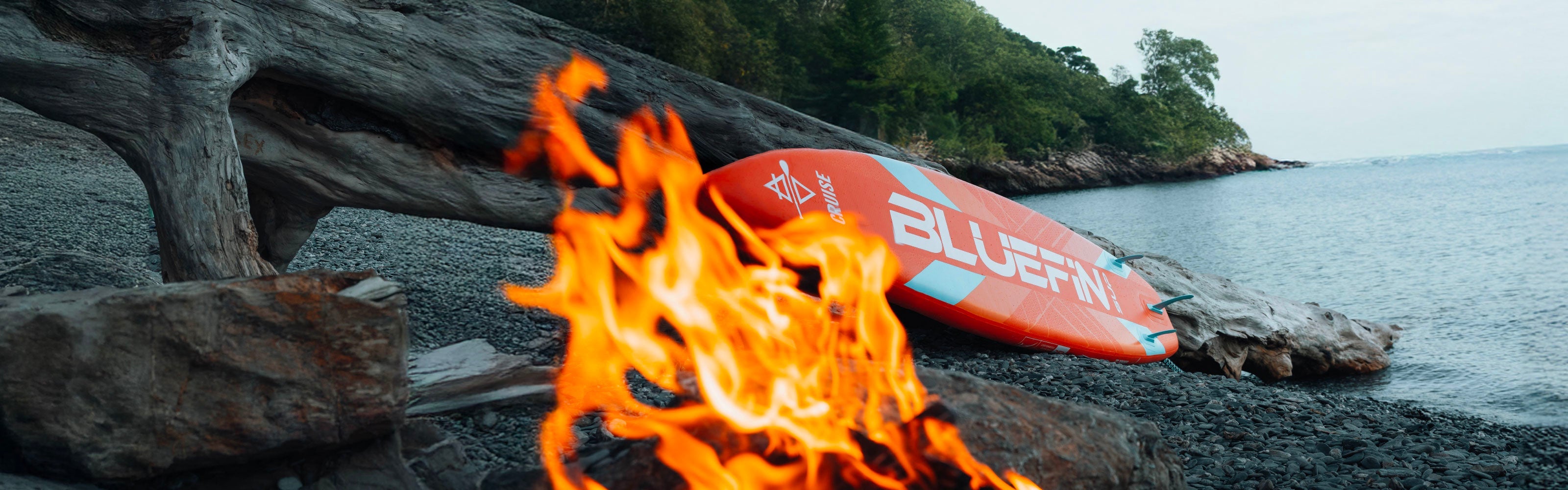 Inflatable paddleboard on rocky shore with fire in foreground and trees in background