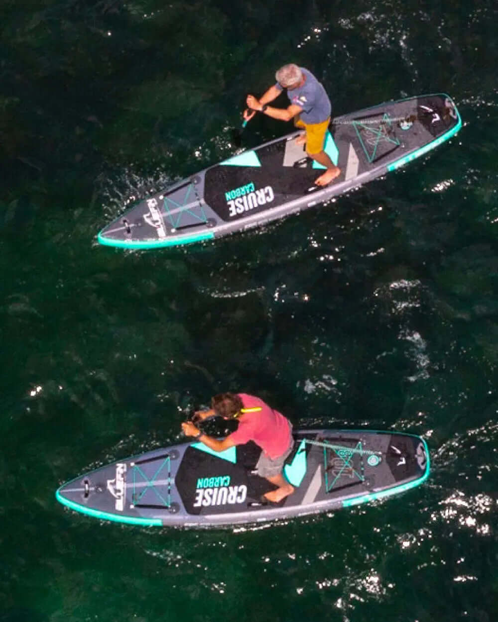 Two people paddleboarding on inflatable boards in clear water, one in yellow shorts and the other in gray shorts.