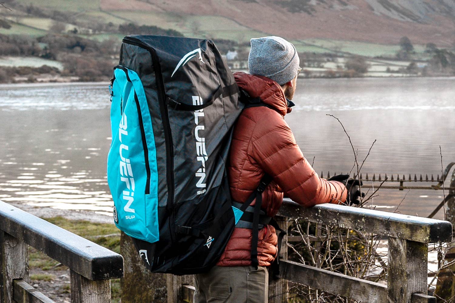 Person in a red jacket with a blue and black backpack overlooking a lake from a wooden railing