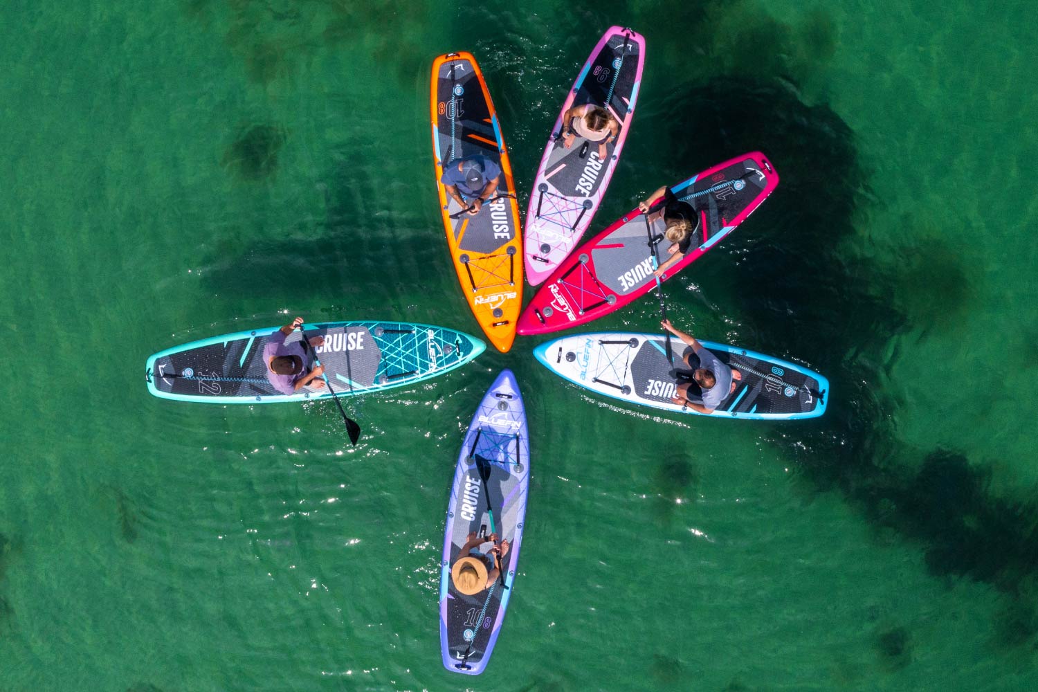 Seis tablas de paddle surf coloridas dispuestas en círculo sobre agua clara, con remeros usando remos.