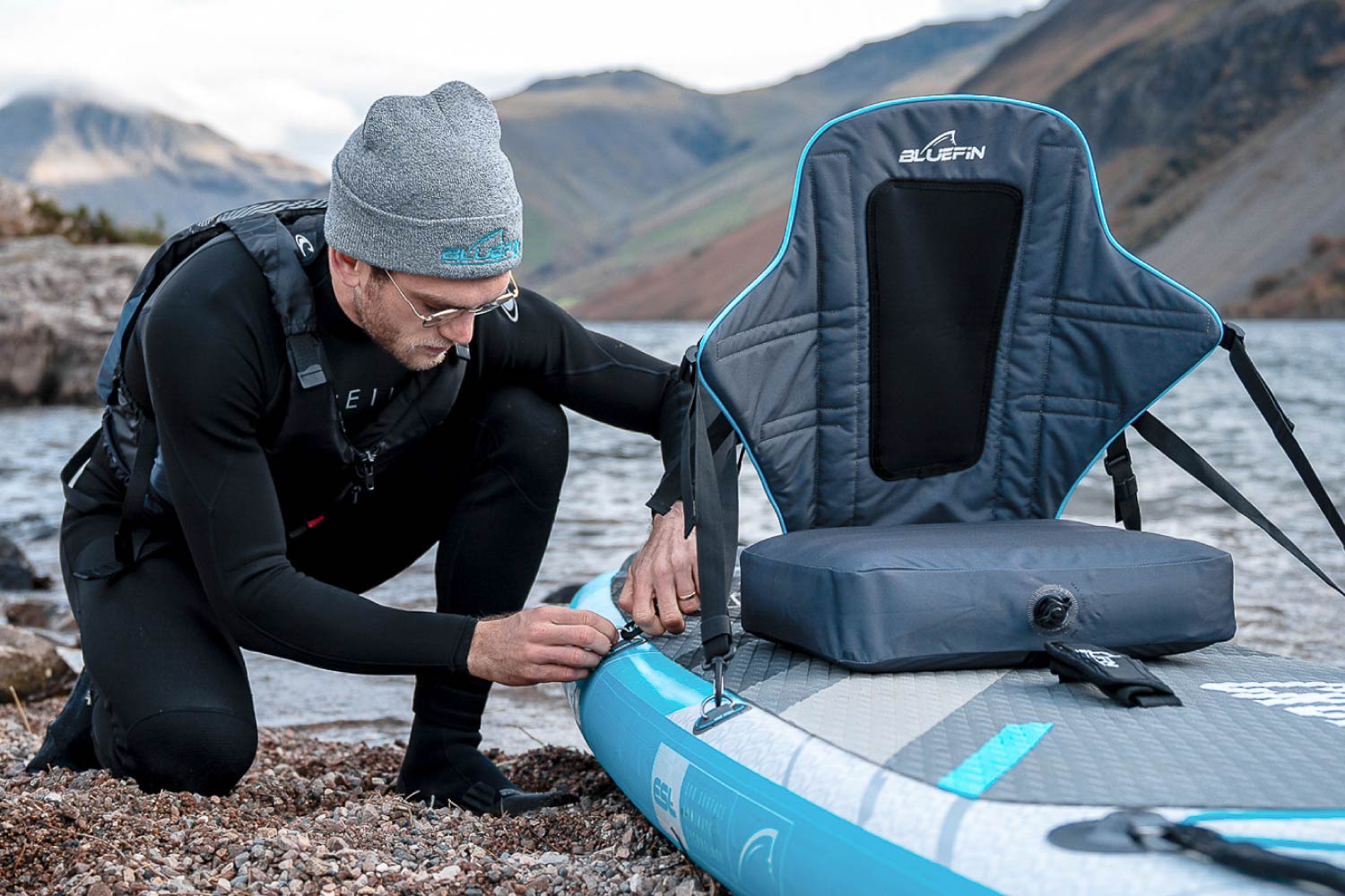 Person in wetsuit adjusting a strap on a Bluefin inflatable paddleboard by the water.