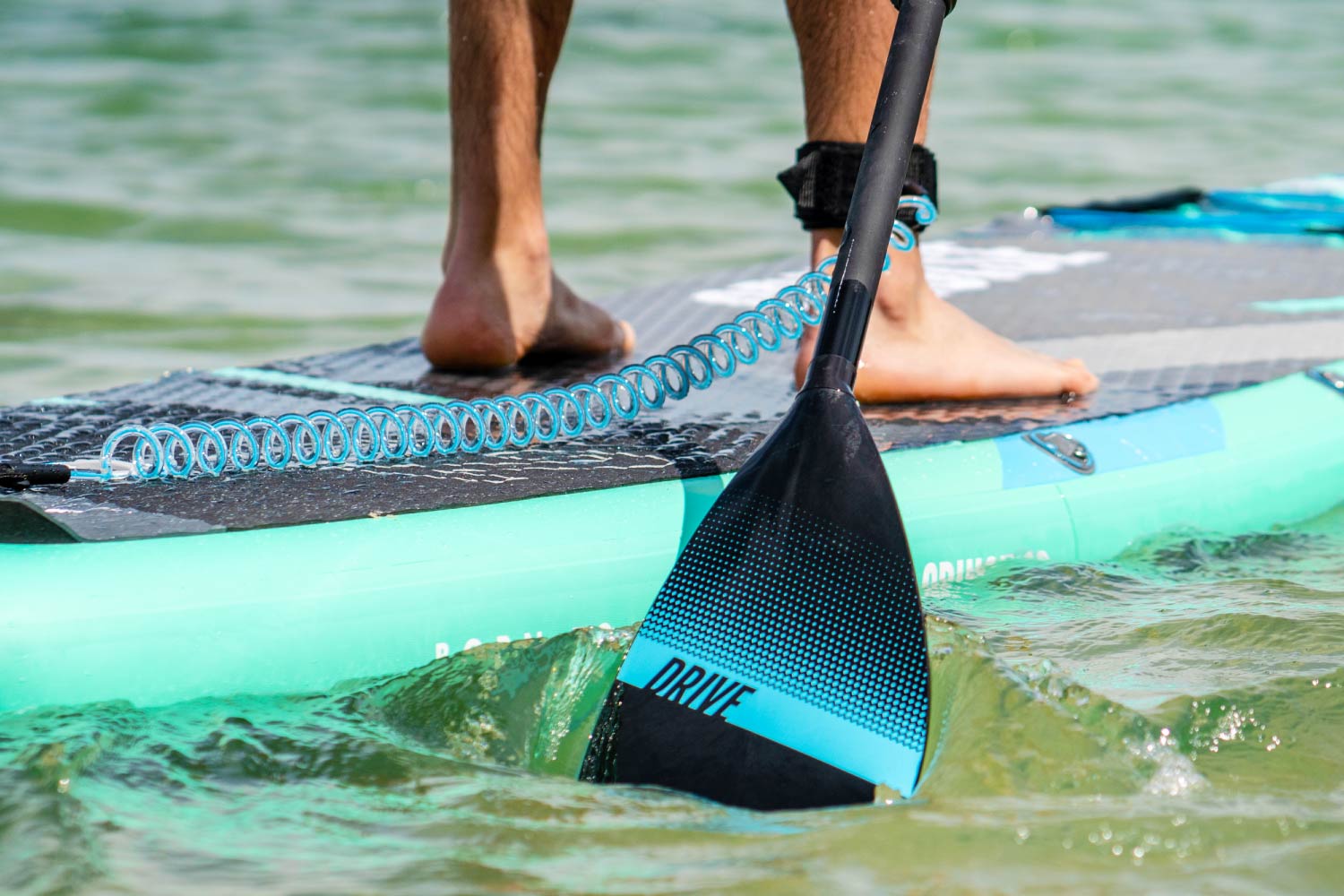 Persona de pie sobre una tabla de paddle surf inflable verde azulado, usando un remo negro con acentos azules en el agua.