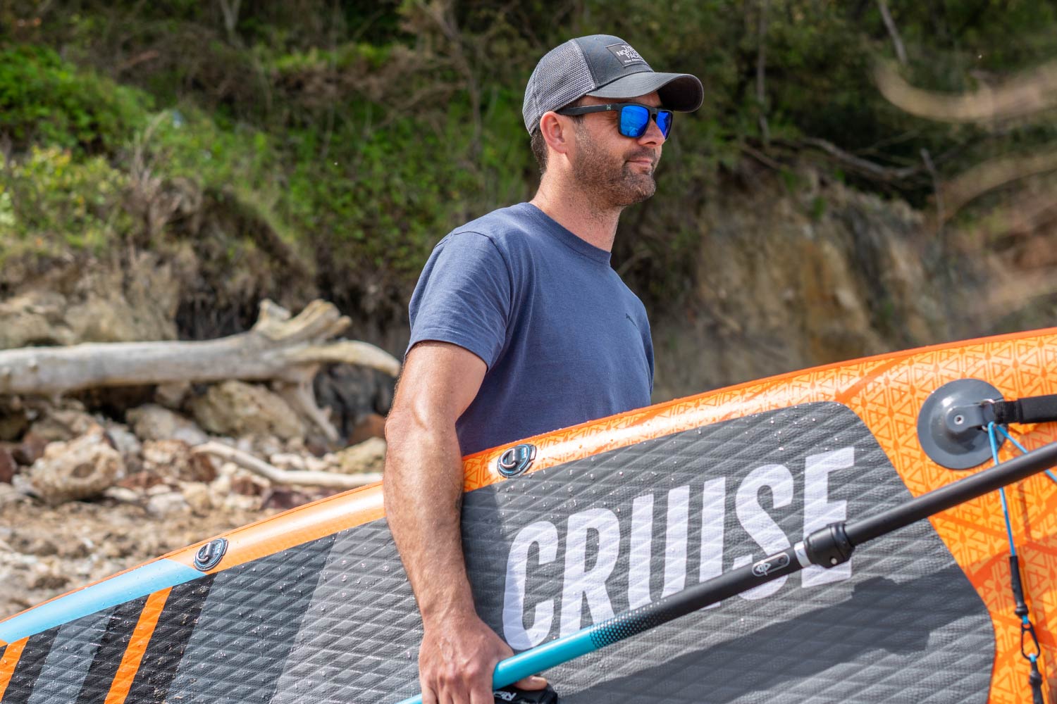 Man holding an inflatable paddleboard with "CRUISE" text, wearing sunglasses and a cap on a beach.