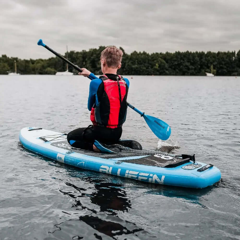 Persona remando en una tabla de paddle inflable azul con una almohadilla de cubierta texturizada en agua tranquila