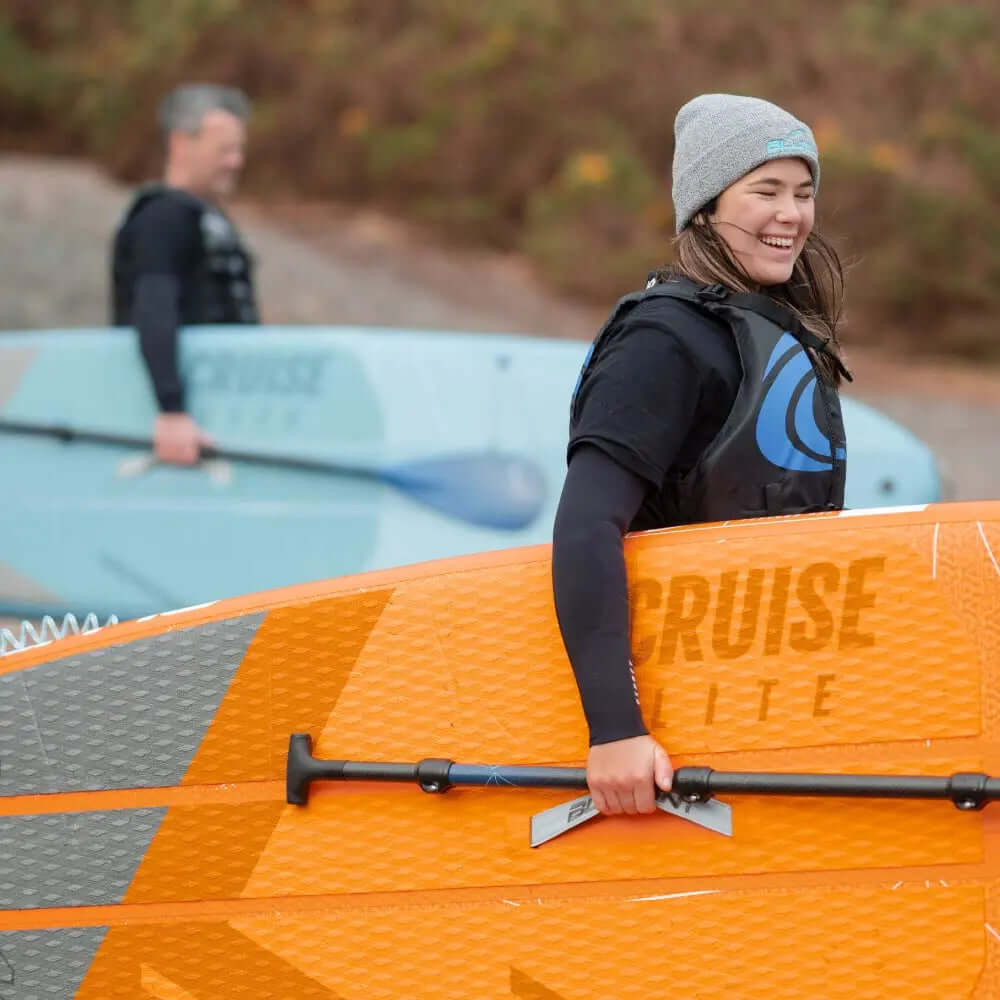 Dos personas en la playa con una tabla de paddle naranja y una tabla de paddle azul claro, sonriendo y preparándose para remar.