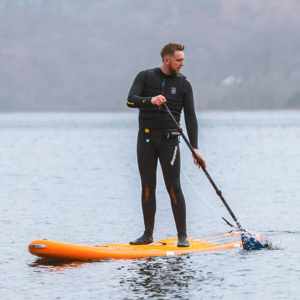 Hombre en traje de neopreno remando en una tabla de paddle de pie naranja en agua tranquila.