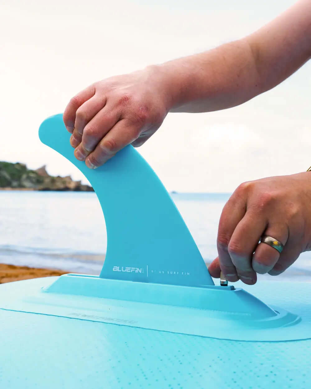 Persona colocando una aleta azul en un paddleboard en una playa con agua de fondo.