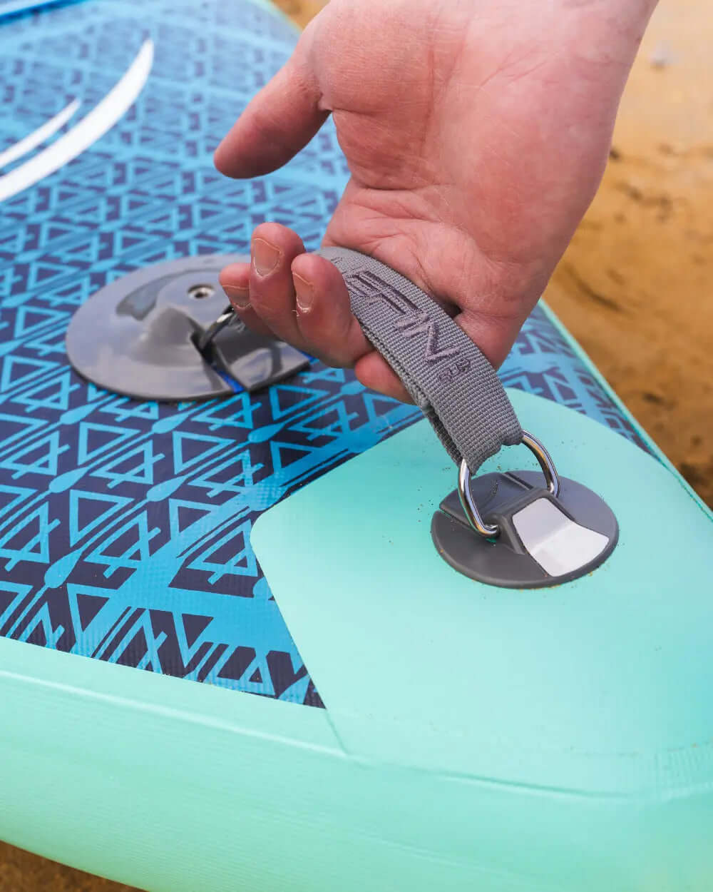 Mano agarrando una correa gris en un paddleboard inflable azul y verde con superficie texturizada.