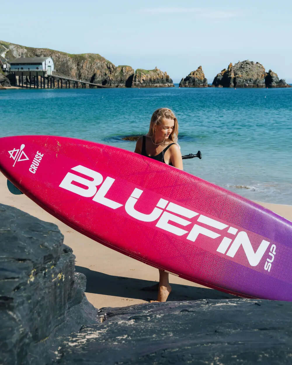 Woman carrying a pink Bluefin SUP paddleboard on a sandy beach with rocky coastline and clear water.
