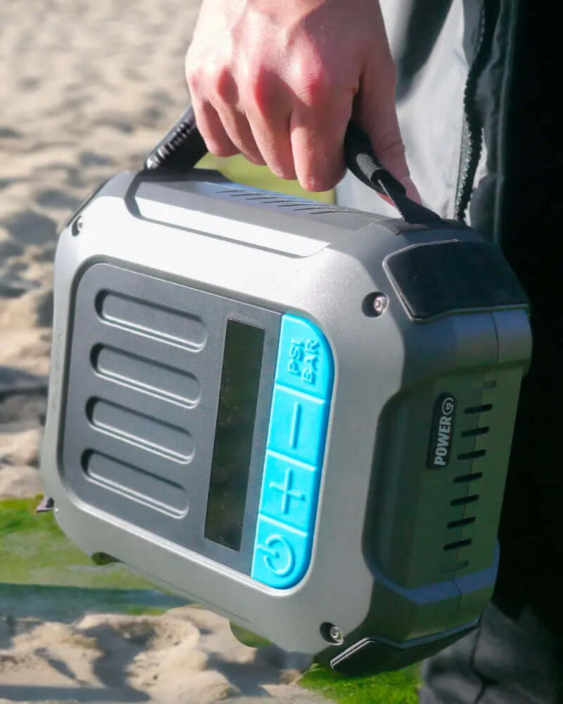 Bomba de aire portátil con panel de control azul, sostenida en una playa de arena.