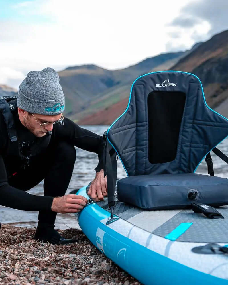 Person in wetsuit adjusting seat on a Bluefin inflatable paddleboard by the water.