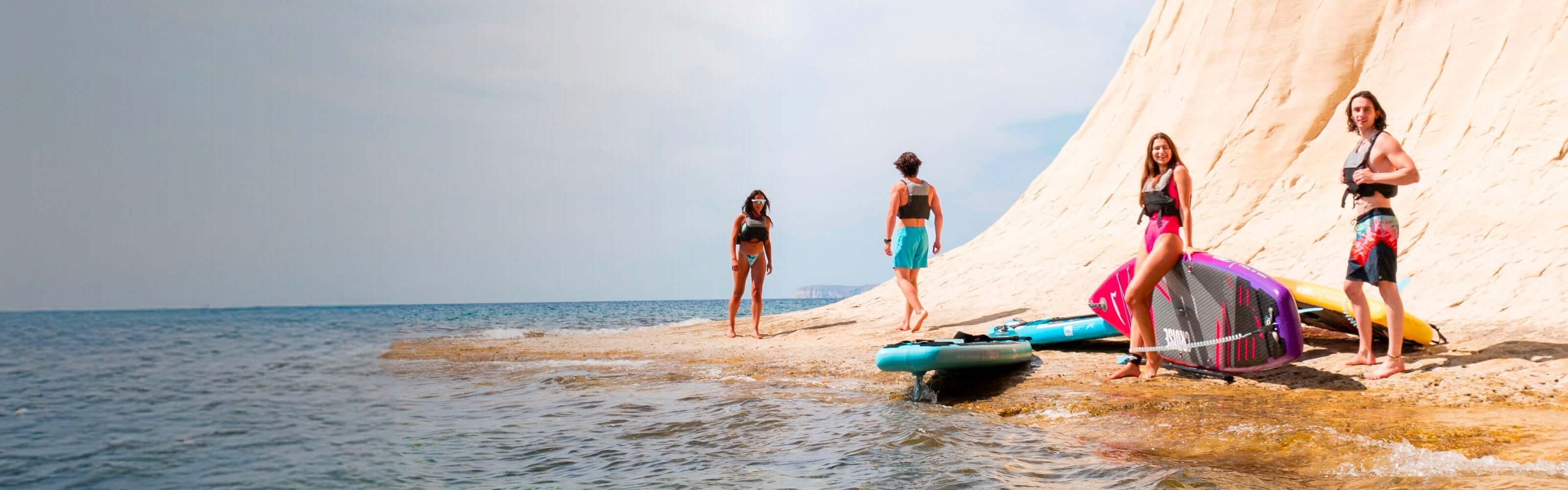 Four people on a beach with paddleboards, wearing swimsuits and life jackets, near a rocky cliff.