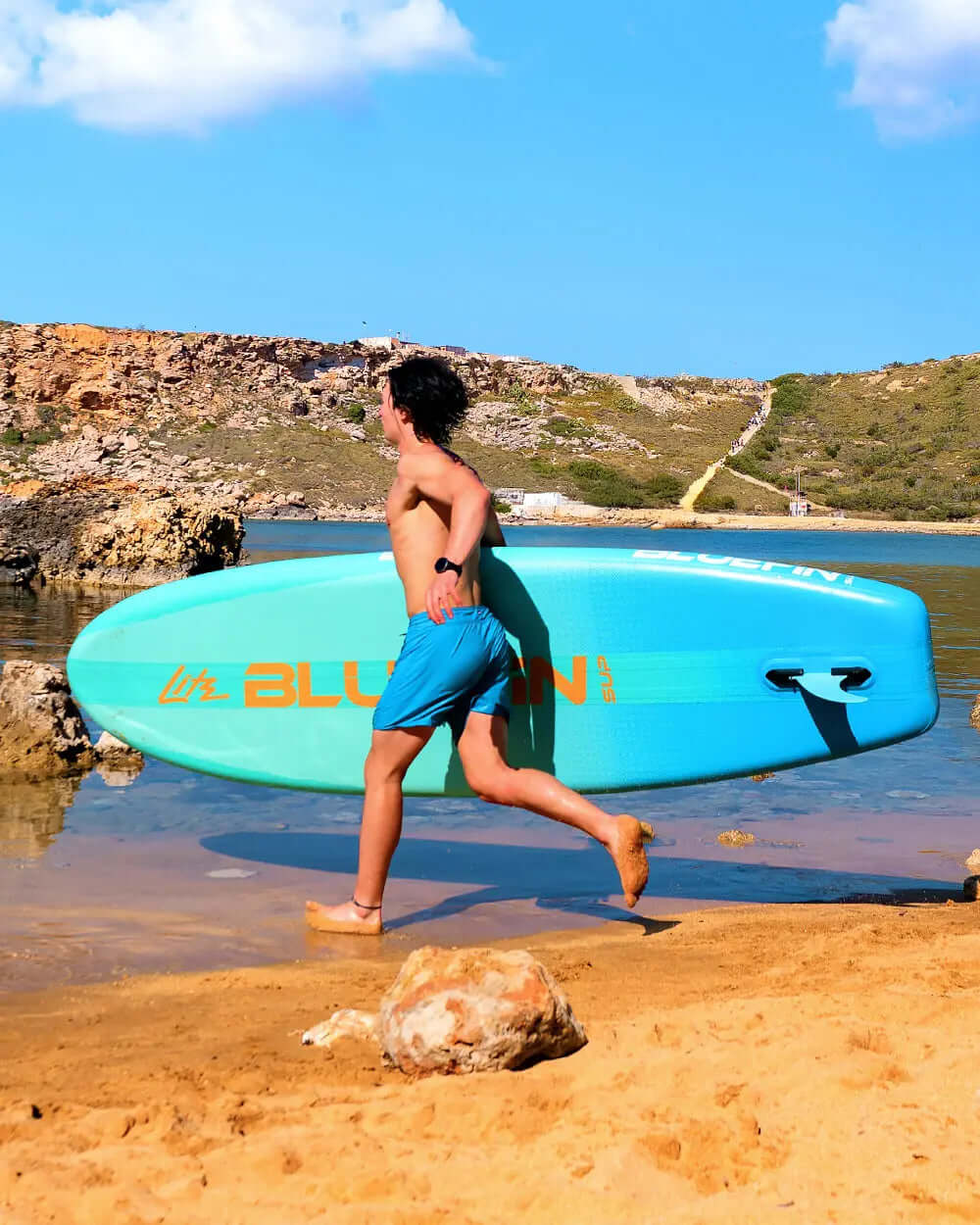 Person running on sand carrying a blue inflatable paddleboard with orange branding and rocky background
