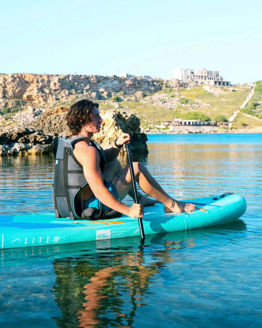 Person sitting on a teal inflatable paddleboard, paddling in calm water with rocky shore in background