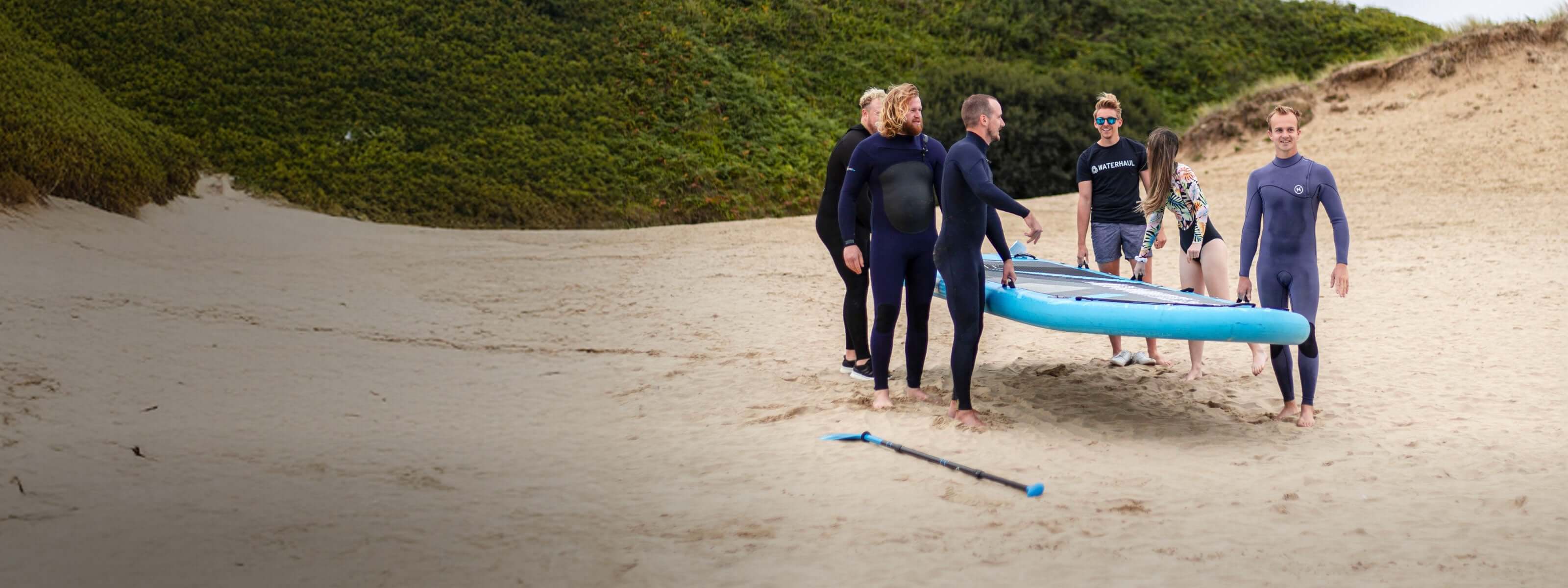 Group of six people in wetsuits carrying a blue paddleboard on sandy beach with greenery in background