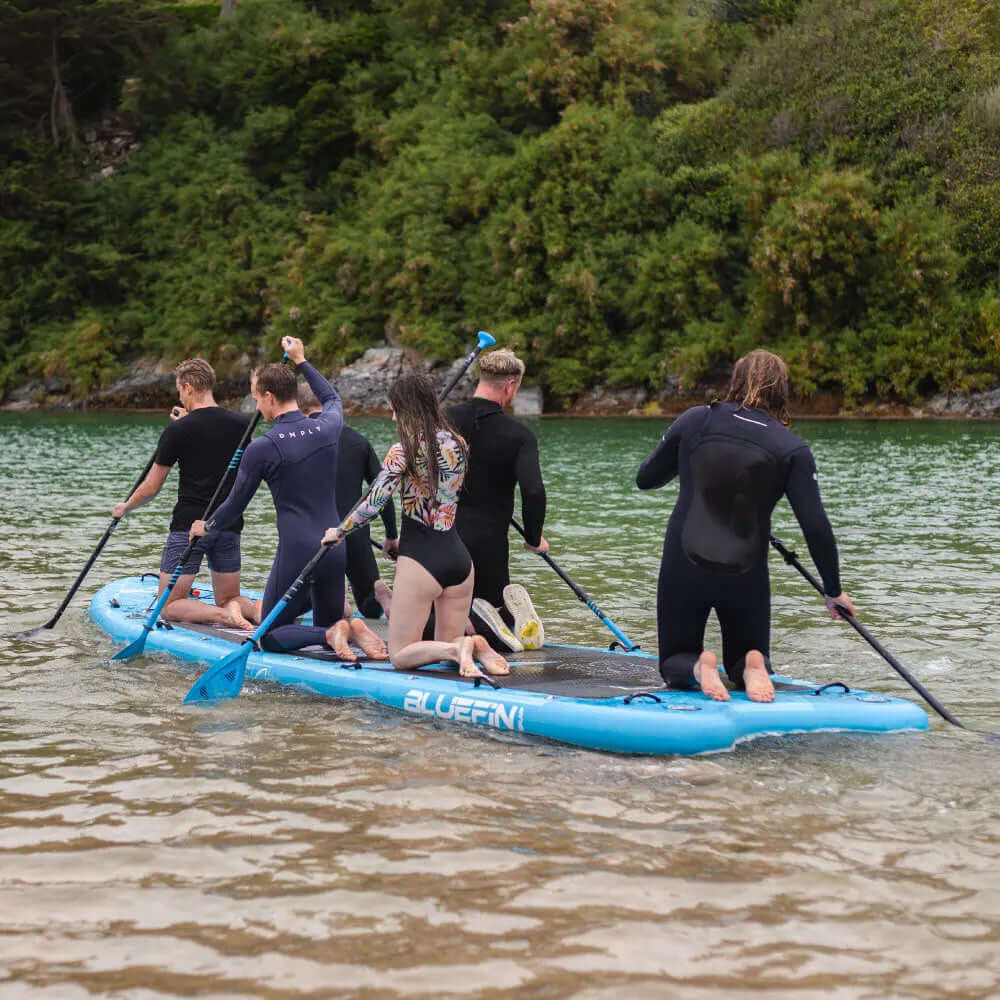 Grupo de seis personas remando en un paddleboard inflable azul en agua tranquila.