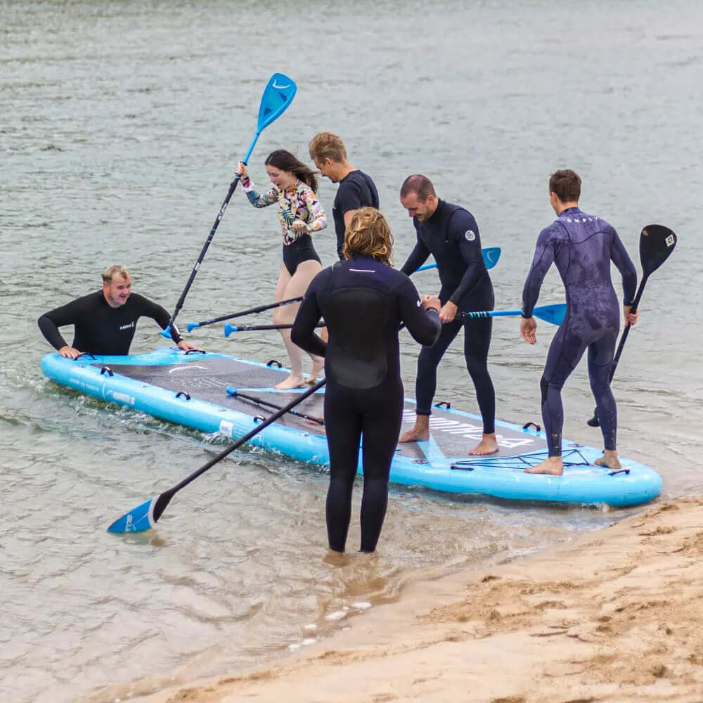 Grupo de seis personas en un gran paddleboard inflable azul en agua poco profunda, algunos remando, otros equilibrándose.