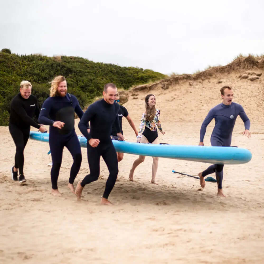 Grupo de seis personas en trajes de neopreno corriendo sobre la arena con una gran tabla de paddle azul