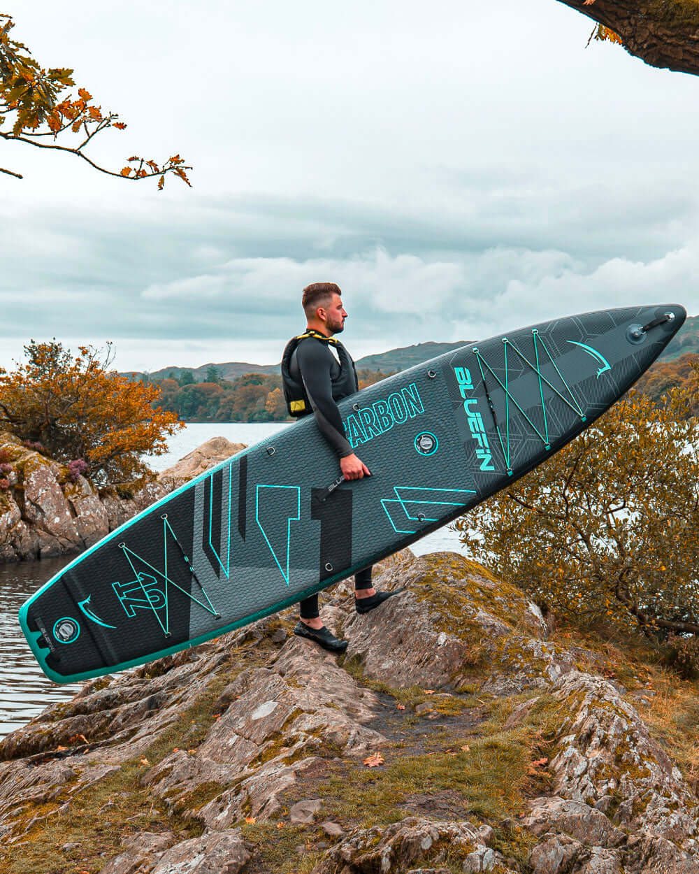 Man in wetsuit carrying a Bluefin Carbon paddleboard by a lake, surrounded by autumn foliage.