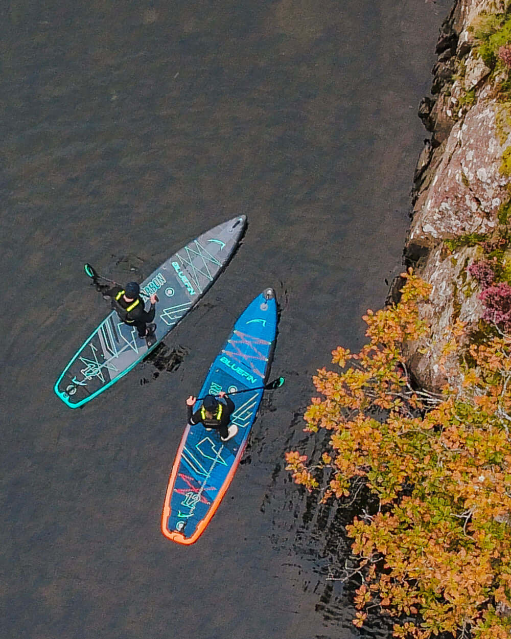 Two paddleboarders on colorful inflatable boards in calm water near a rocky shore with autumn foliage.