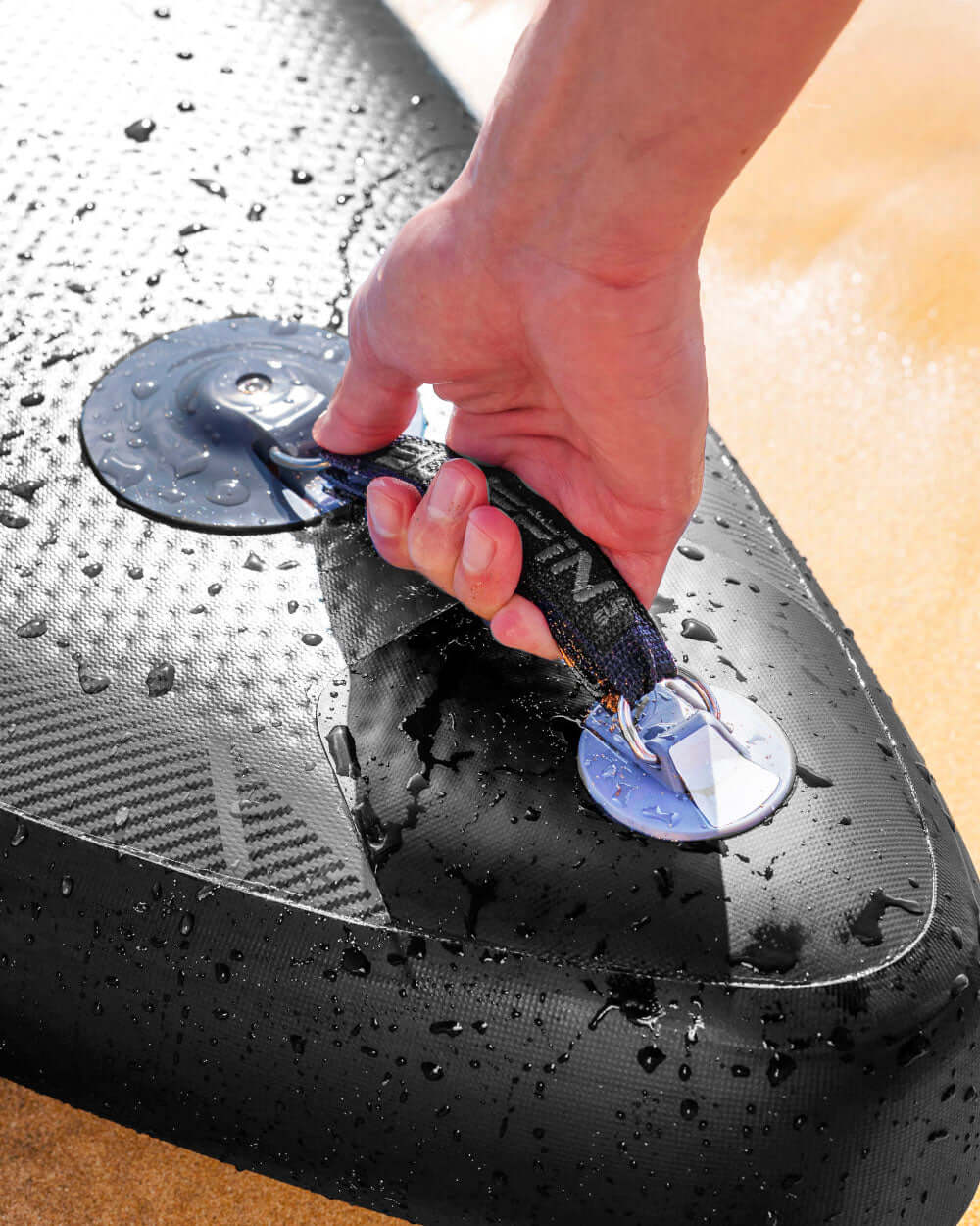 Hand gripping a black paddleboard handle with water droplets on the surface and sandy background