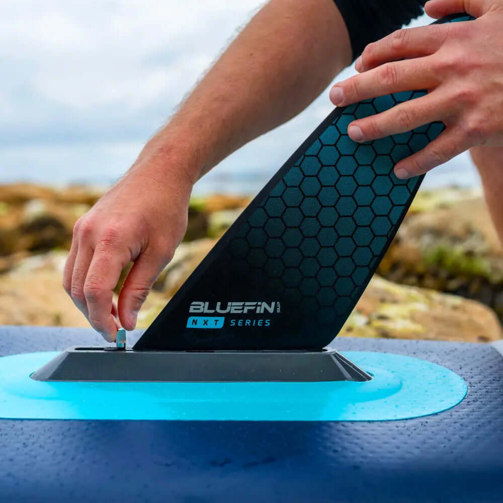 Persona colocando una aleta hexagonal negra en una tabla de paddle Bluefin en una costa rocosa.