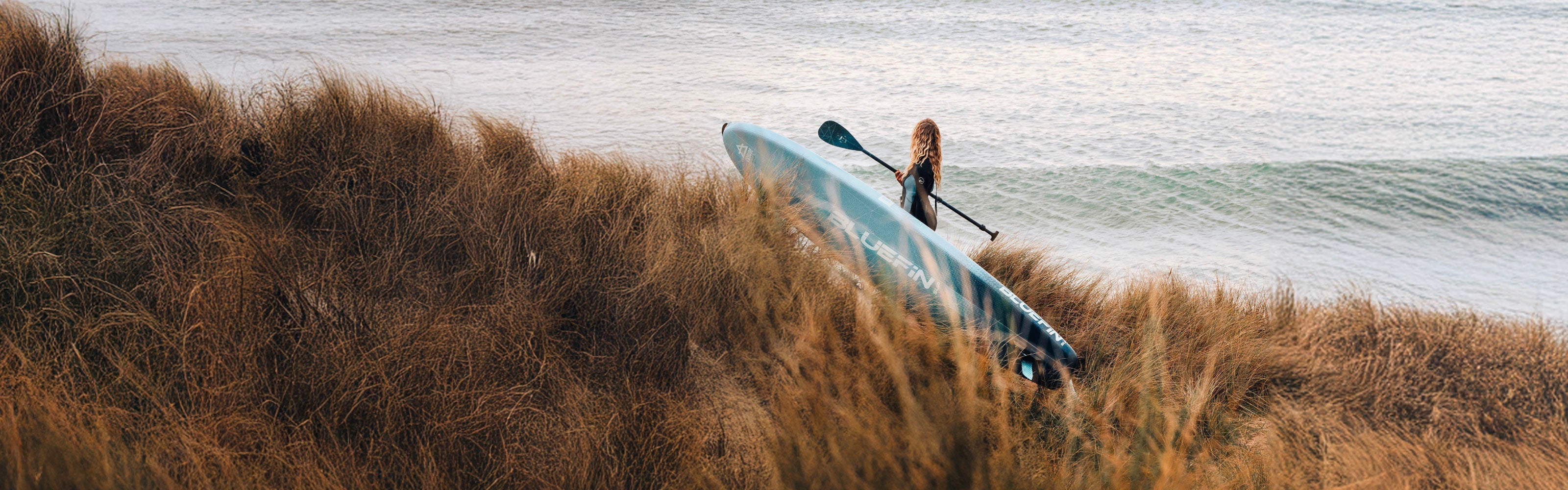 Person carrying a paddleboard through tall grass towards the water, with gentle waves in the background.
