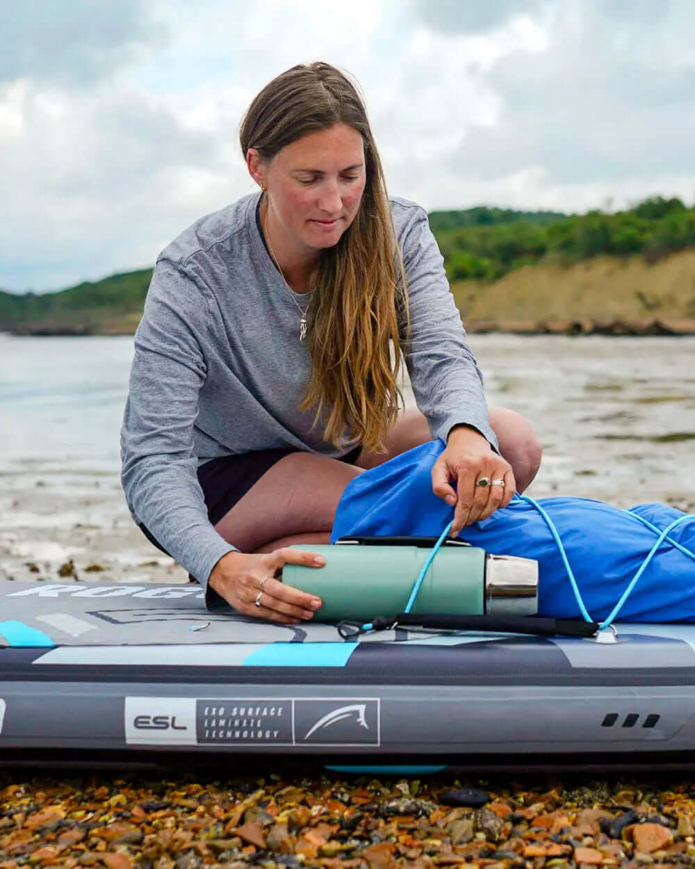 Person securing a green water bottle on a blue bag while sitting on an inflatable paddleboard by the shore.
