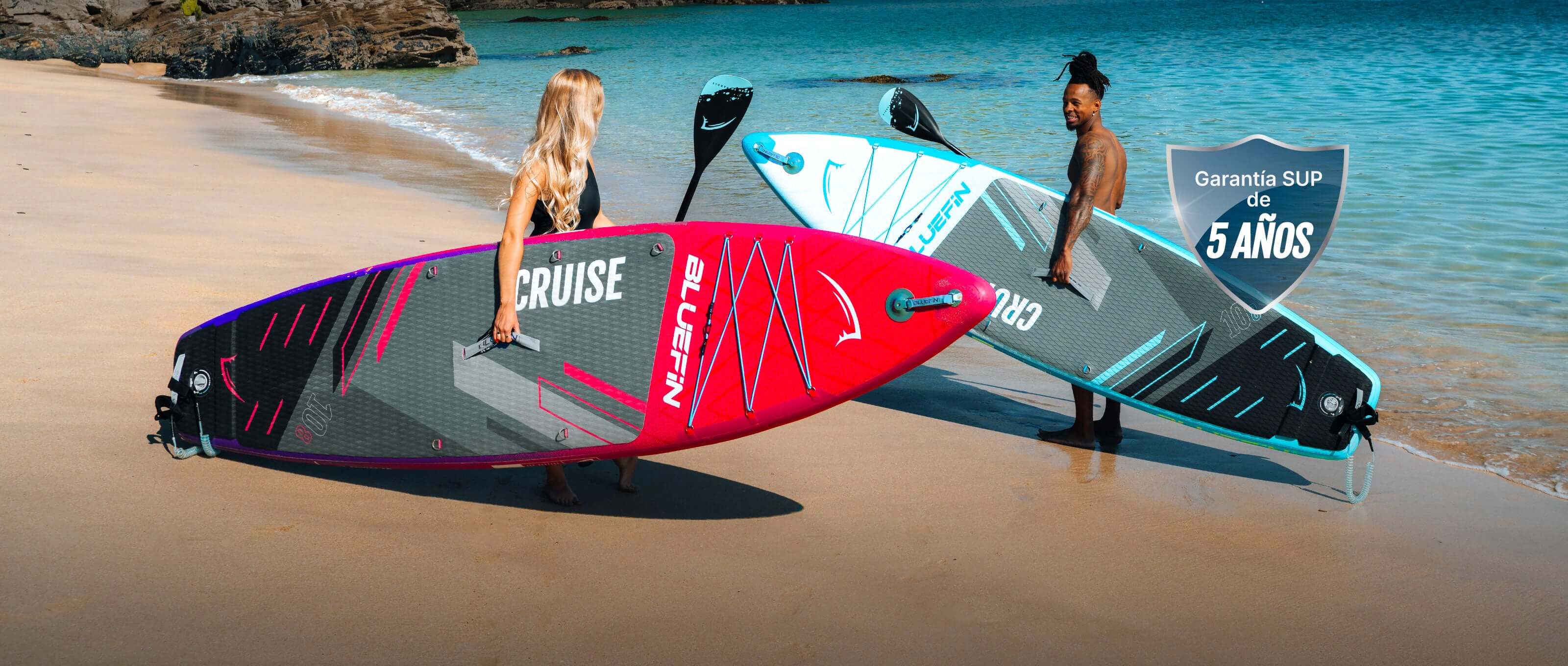 Two inflatable paddleboards on a beach, one pink and one blue, with paddles and ocean in background.