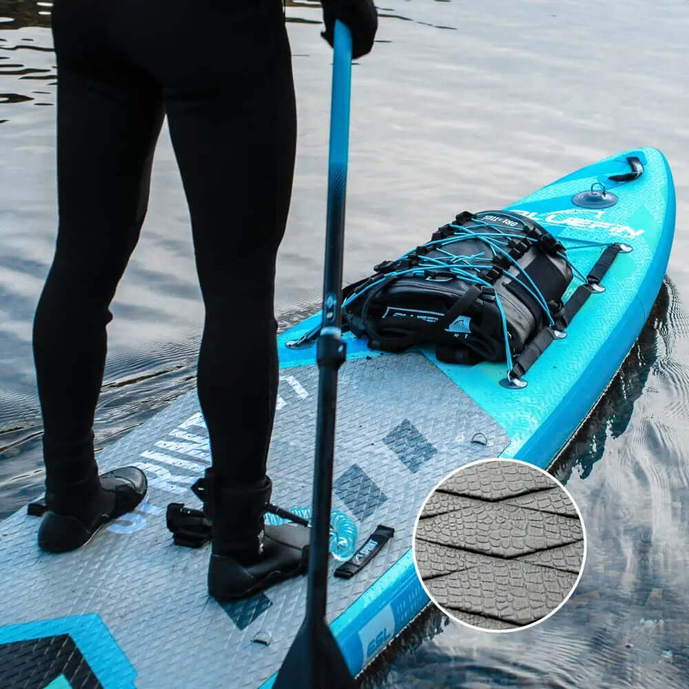 Tabla de paddle con pad de cubierta texturizado, mochila negra asegurada, persona sosteniendo un remo en el agua