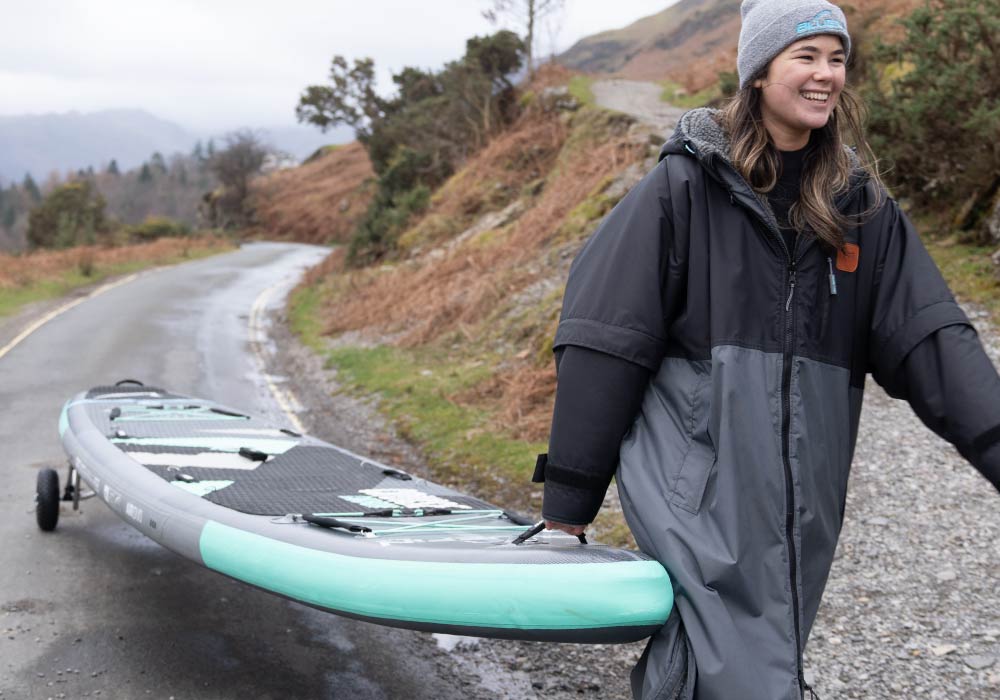 Person in a black and gray jacket carrying a paddleboard along a road with hills in the background