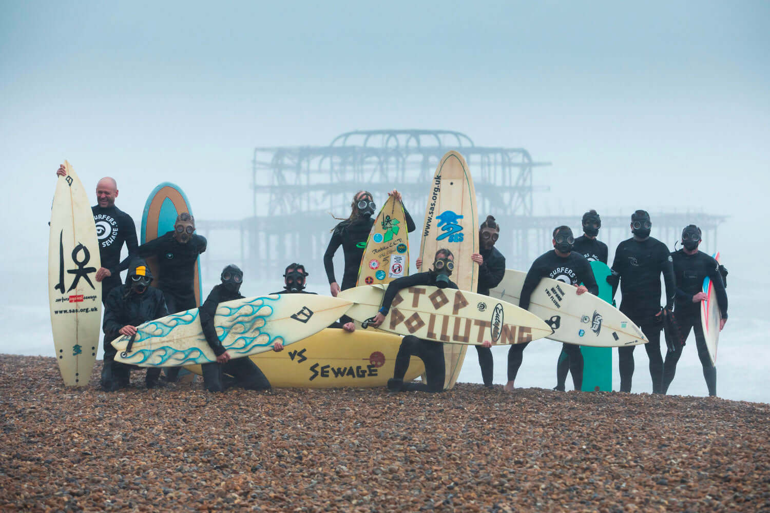 Group of surfers in wetsuits and gas masks holding surfboards on a pebbly beach with a pier in the background.