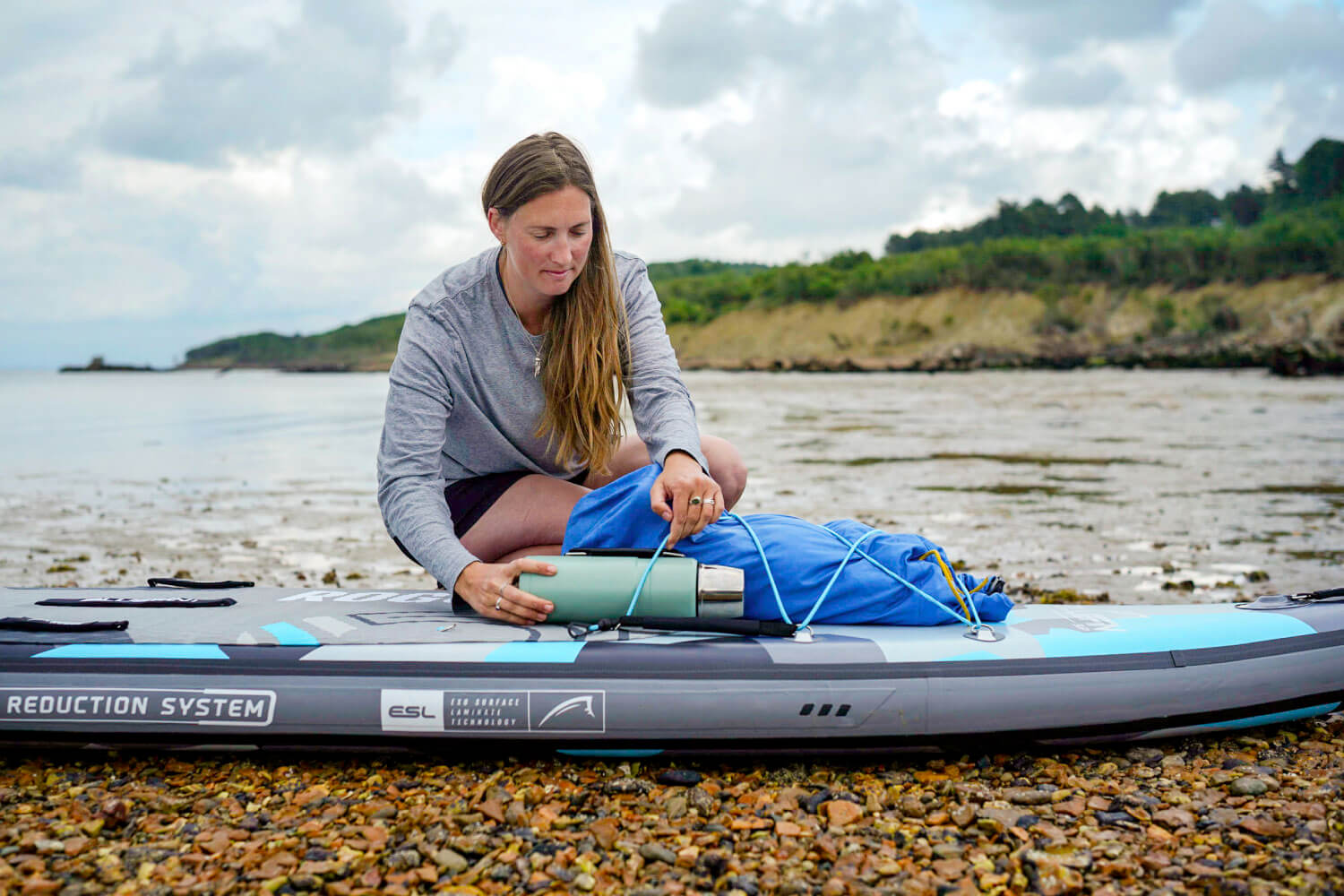 Woman securing a blue bag and thermos on an inflatable paddleboard by the shore