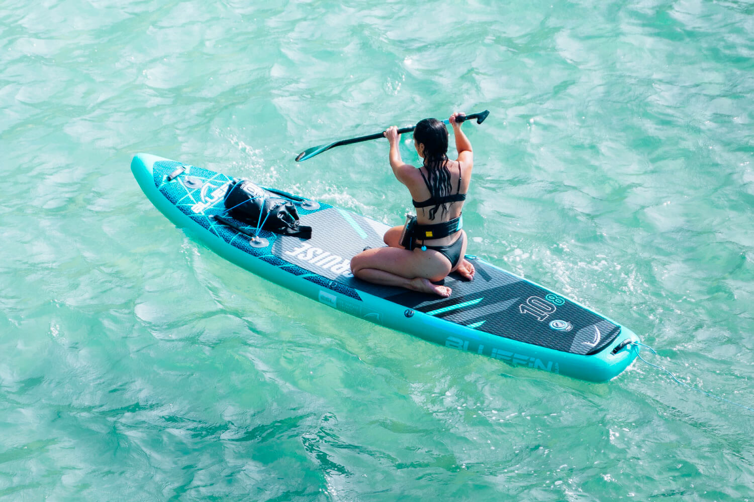 Person paddling on a turquoise inflatable paddleboard in clear water, wearing a black bikini.