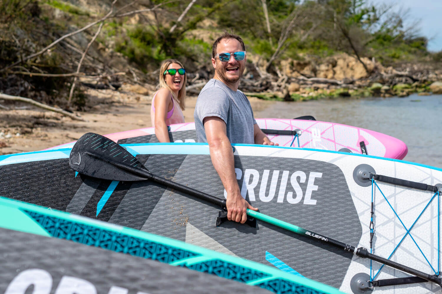 Two people smiling near inflatable paddleboards on a beach, one holding a paddle.