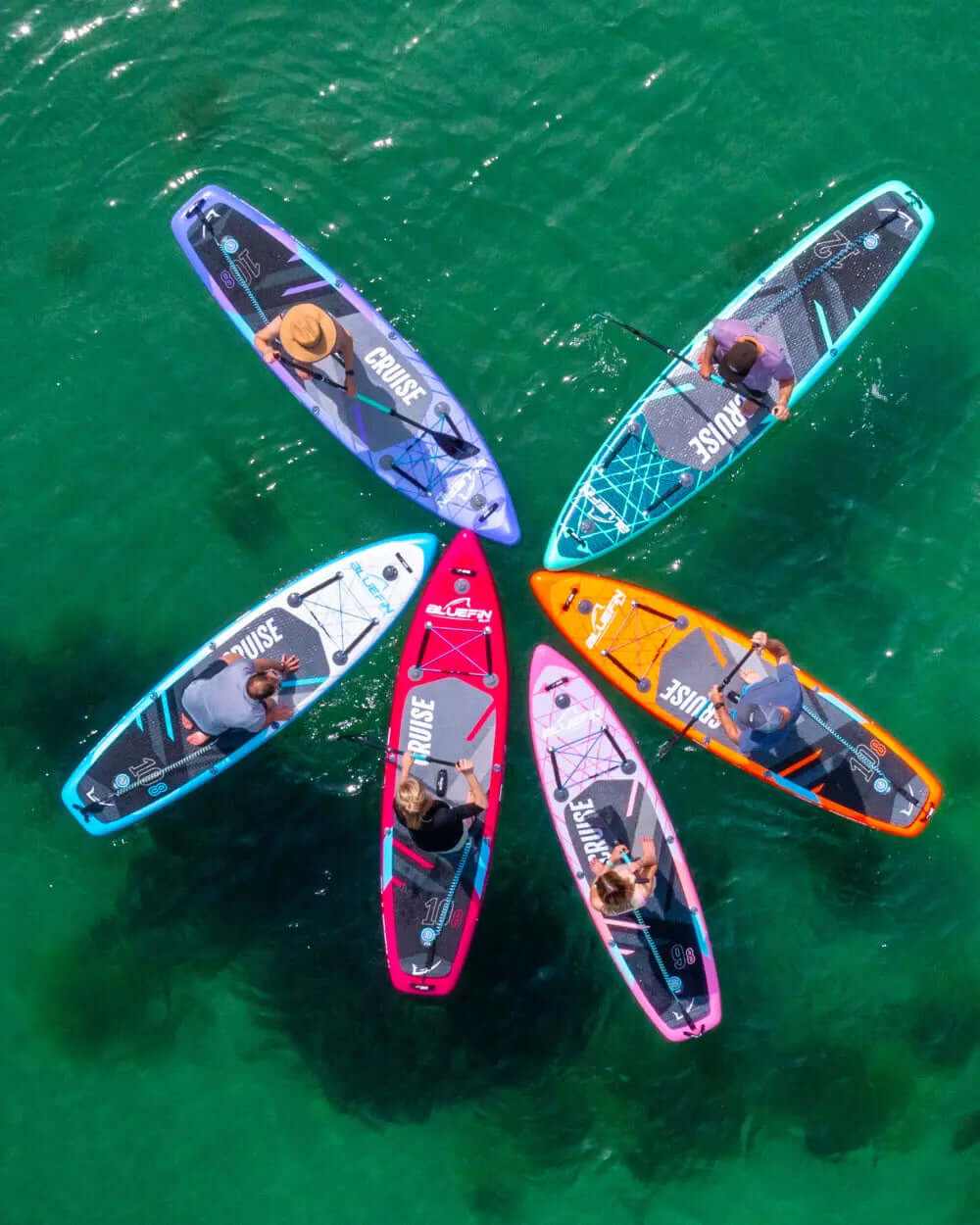 Six colorful paddleboards arranged in a circle on clear water, with paddlers visible on each board.