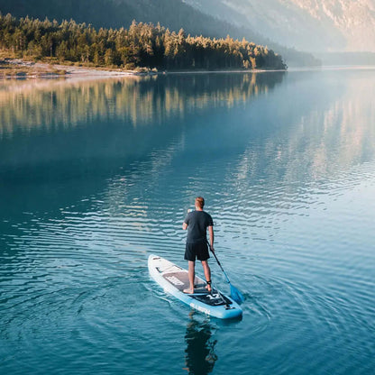 Persona remando en una tabla de paddle surf azul en aguas tranquilas con montañas boscosas al fondo
