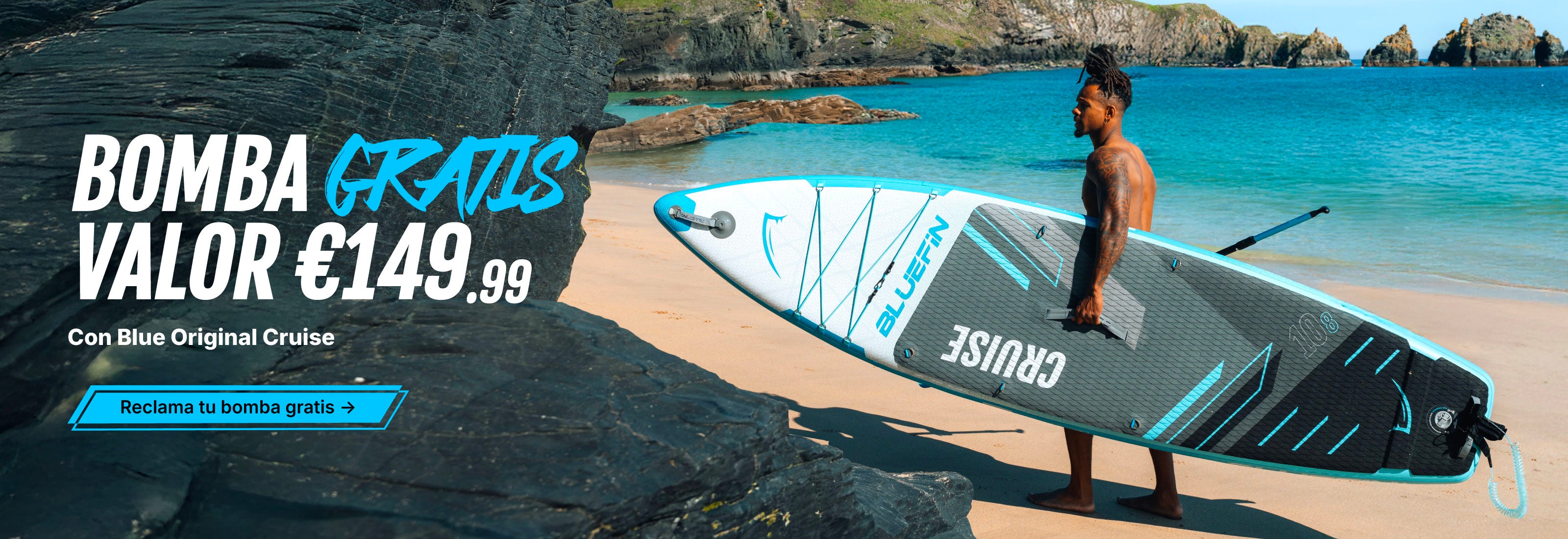Man carrying a Bluefin inflatable paddleboard on a beach with rocky coastline and clear water.