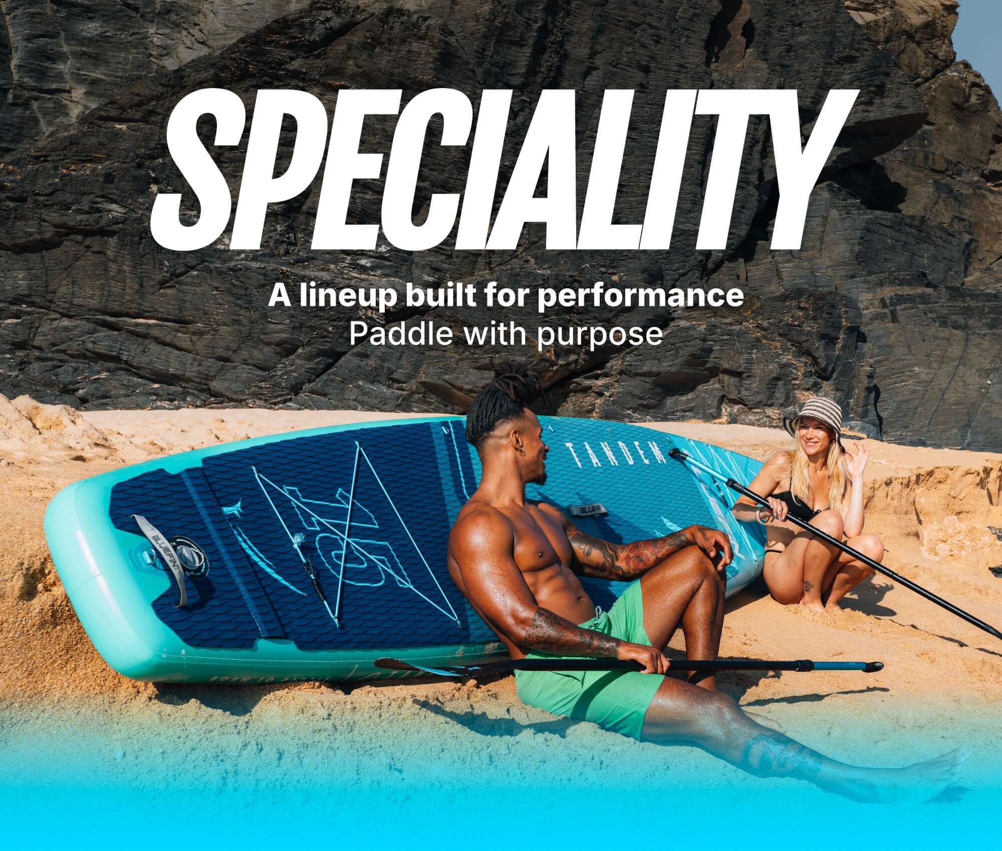 Two people on a beach with an inflatable paddleboard, one sitting and one kneeling, both holding paddles.