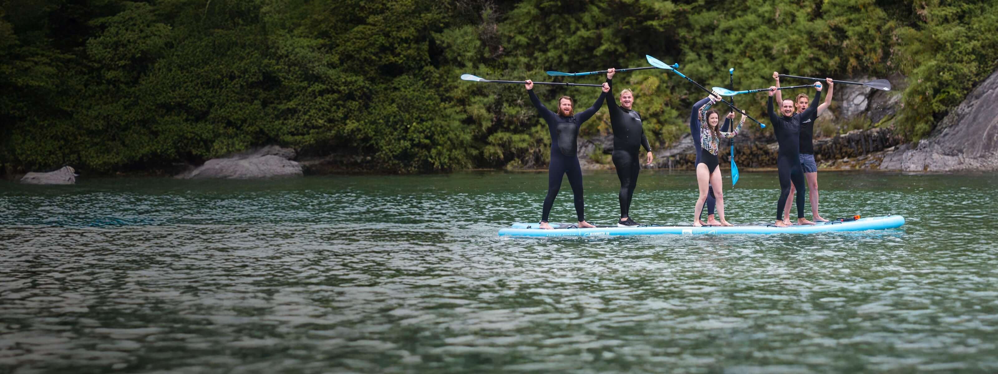 Group of five people on a paddleboard, smiling and holding paddles above their heads on calm water.