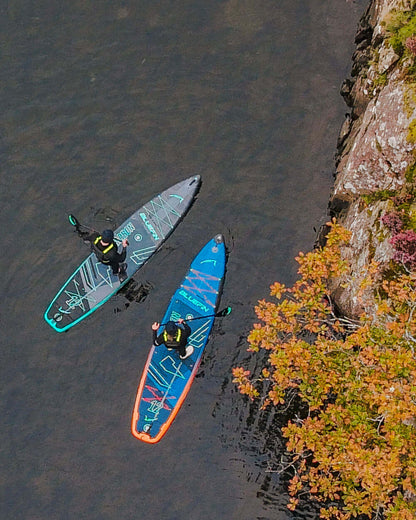 Two paddleboarders on colorful inflatable boards in calm water near a rocky shore with autumn foliage.