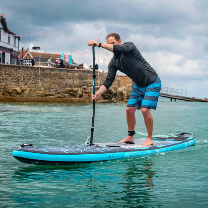 Hombre remando en una tabla de paddle inflable azul en agua tranquila, vistiendo camiseta negra y pantalones cortos a rayas.
