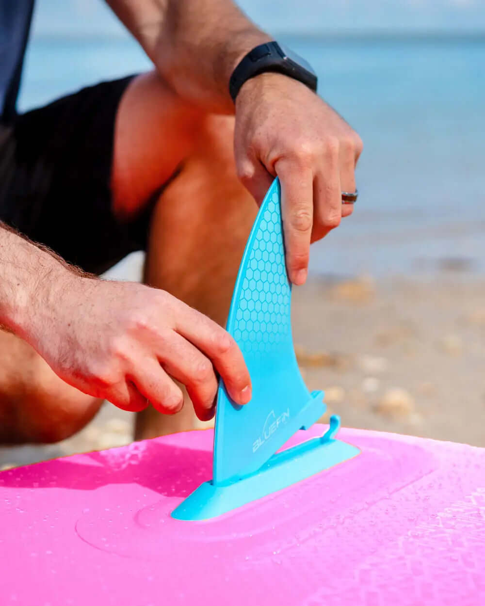Person attaching a blue fin to a pink inflatable paddleboard on a beach