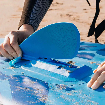 Persona uniendo una aleta azul a una tabla de paddle inflable en una playa de arena.