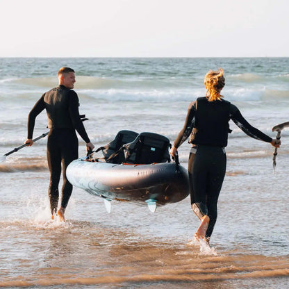 Dos personas en trajes de neopreno llevando una tabla de paddle inflable a través de aguas poco profundas en la playa