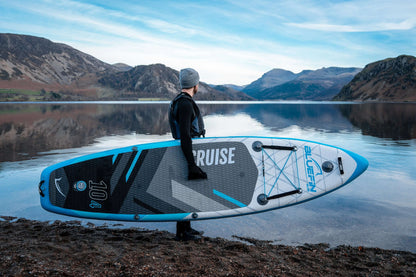 Person in wetsuit holding a Bluefin SUP paddleboard by a calm lake with mountains in the background